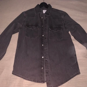 Svart jeansskjorta med tryckknappar - Säljer en svart jeansskjorta  i storlek L. Skjortan har två bröstfickor med lock och tryckknappar framtill. Den är långärmad och har en klassisk krage, perfekt för en avslappnad och cool look. Aldrig andvänd. 