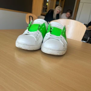 Valentino Garavani Open sneakers med grönt band - Säljer ett par vita Valentino Garavani Open sneakers med ljus grönt i mitten.  Skorna har rund tå, platt sula och klassisk snörning. Perfekta för dig som vill sticka ut med en lyxig touch, passar 41, priset är inte hugget i sten 