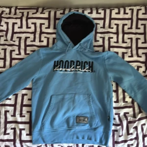 Blå hoodie från Hoodrich  - Snygg ljusblå hoodie från Hoodrich med broderad logga i svart och vitt på bröstet. Tröjan har huva med snörning, känguruficka och en liten patch nertill. Perfekt för dig som gillar streetwear och vill sticka ut lite extra.