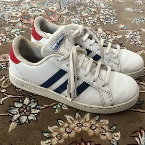 Vita adidas sneakers med blå ränder - Snygga vita adidas sneakers med klassiska blå ränder på sidorna och röd detalj på hälen. Skorna har snörning och platt sula, samt adidas-logga på plös och häl. Perfekta för en sportig och stilren look.Priset kan deskutera.