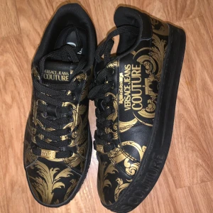 Svarta och guldiga Versace Jeans Couture sneakers - Snygga sneakers från Versace Jeans Couture i svart med lyxigt guldigt barockmönster. Skorna har snörning, rund tå och platt sula med logga på sidan och sulan. Perfekta för dig som vill sticka ut med en exklusiv vibe.”Dem säljs inte mer ute på någon hemsida”. Skriv för frågor