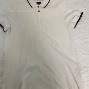 Vit piké från Armani Exchange XL - Vit pikétröja från Armani Exchange i storlek XL. Klassisk design med korta ärmar, krage och knappar framtill. Röd och svart randig detalj på krage och ärmslut. Tillverkad i mjuk bomull för en clean och stilren look.