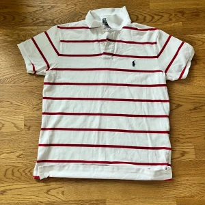 randig pikétröja Polo Ralph Lauren - Vit pikétröja från Polo Ralph Lauren med röda horisontella ränder och klassisk krage. Tröjan har korta ärmar, knappar vid halsen och den ikoniska broderade loggan på bröstet. Perfekt för en clean och sportig stil.