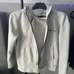 Ljusgrå hoodie från Sail Racing - Ljusgrå hoodie från Sail Racing med dragkedja framtill och två fickor. Mjuk insida och huva med dragsko. Diskret logga på bröstet. Perfekt för en avslappnad stil.