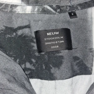 Svartvit skjorta med palmmotiv från Neuw - Säljer en svartvit skjorta från Neuw med tryckt palmmotiv. Skjortan har korta ärmar, klassisk krage och knäppning framtill. Perfekt för dig som gillar unika mönster och vill sticka ut lite extra.