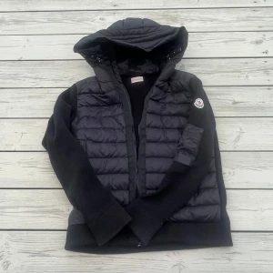 Moncler Cardigan - Säljer en svart Moncler-jacka med stickade ärmar och quiltad front. Jackan har huva, dragkedja och Moncler-logga på ärmen. Perfekt för lager-på-lager och snygg till vardags. Självklart äkta och Scan fungerar
