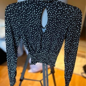 Svart prickig blus med lång ärm och knytning i nacken - Svart blus med vita prickar, långärmad och med smockad midja. Blusen har en snygg öppning framtill vid halsen och knytning i nacken. Perfekt för dig som gillar trendiga detaljer och mönster.