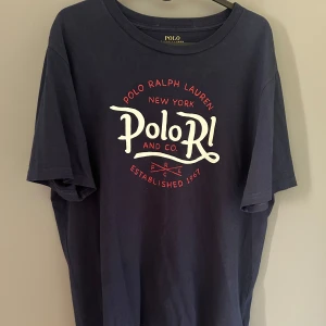 Mörkblå t-shirt från Polo Ralph Lauren - Mörkblå t-shirt från Polo Ralph Lauren med vit och röd tryckt logga framtill. Klassisk rund halsringning och normal passform. Perfekt för en avslappnad och stilren look. Storlek XL men passar M/L