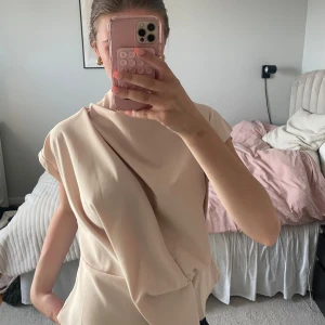 Beige omlott topp med drapering - Säljer en beige omlott topp med snygg drapering och asymmetrisk design. Toppen är ärmlös och har en elegant, stilren look som passar perfekt till många olika outfits.