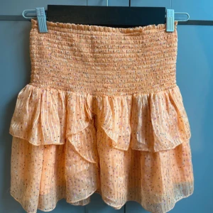 Persikofärgad volangkjol med glitter - Supersöt persikofärgad kjol med smock i midjan och volanger. Kjolen har tunna glittriga ränder och små prickar i olika färger. Perfekt för dig som vill ha en lekfull och trendig look.