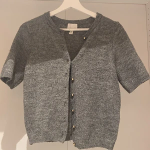 Grå stickad kofta med guld knappar  - Kortärmad grå stickad kofta från H&M i storlek XS. Som har guldknappar. 