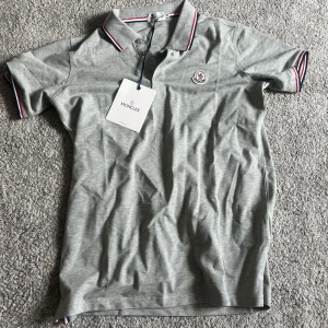 Grå piké t-shirt från Moncler - Snygg grå piké t-shirt från Moncler med klassisk krage och knappar. Tunn röd, vit och blå rand på krage och ärmslut. Moncler-logga broderad på bröstet. Perfekt för en stilren och sportig look. Skriv vid frågor!