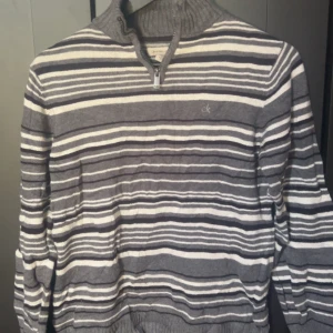 Randig stickad tröja från Calvin Klein Jeans - Säljer en grå och vit randig stickad tröja från Calvin Klein Jeans med halv dragkedja och hög krage. Tröjan har långa ärmar och broderad CK-logga på bröstet. Perfekt för en stilren och avslappnad look.