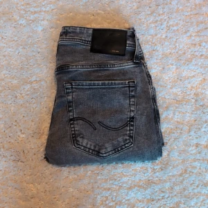Grå slitna jeans från Jack & Jones - Grå jeans från Jack & Jones med snygga slitningar och en tvättad look. Modellen har klassiska fem fickor, bälteshällor och knappgylf. Jeansen är i stretchigt denim och har en smal passform som sitter snyggt längs benen.
