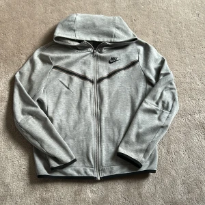 Grå zip-hoodie från Nike - Snygg grå hoodie från Nike med dragkedja och huva. Jackan har svarta detaljer, diskret logga på bröstet och en praktisk ficka med dragkedja på ärmen. Perfekt för en sportig och avslappnad stil. ( pris kan diskuteras)