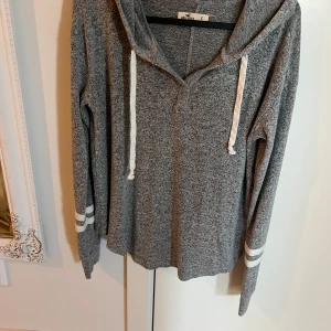 Grå hoodie från Hollister med vita detaljer - Mysig grå hoodie från Hollister med vita snören och dubbla vita ränder på ärmarna. Tröjan har lång ärm, huva och är tillverkad i ett mjukt material som passar perfekt för kyligare dagar.