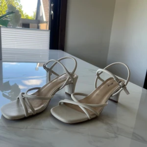 Vita sandaler med klack från Alley - Säljer ett par stilrena vita sandaler från Alley med klack och smala remmar. Skorna har öppen tå, fyrkantig framdel och justerbart spänne vid ankeln. De användes 2 gånger,är då i mycket bra skick och är i storlek 38