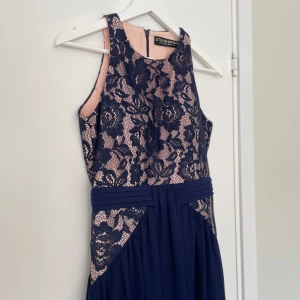 Marinblå långklänning med spets från Little Mistress - Säljer en elegant marinblå långklänning från Little Mistress med vacker spetsdetalj upptill och beige underton. Klänningen är ärmlös och har markerad midja samt plisserad kjol. Perfekt som balklänning, aftonklänning eller festliga tillfällen.