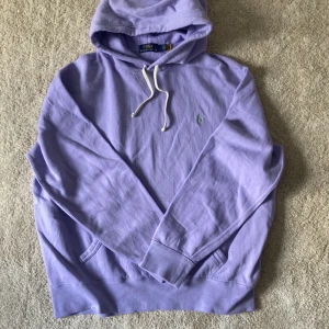 Ljuslila hoodie från Polo Ralph Lauren - Säljer en ljuslila hoodie från Polo Ralph Lauren med klassisk broderad logga på bröstet. Tröjan har huva med vita snören och en stor ficka framtill. Perfekt för en avslappnad stil.