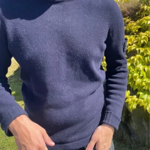 Mörkblå stickad tröja från Stone Island - Säljer en mörkblå stickad tröja från Stone Island med klassisk patch på vänster ärm. Tröjan har rund halsringning och långa ärmar med ribbade muddar. Perfekt för kyligare dagar och enkel att matcha med olika outfits.