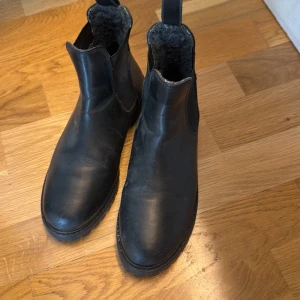 Svarta chelsea boots med ullfoder - Svarta chelsea boots från Furlin med mjukt ullfoder och elastiska paneler på sidorna. Skorna har rund tå och platt sula, perfekta för kyligare dagar. Klassisk och stilren design som passar till mycket.