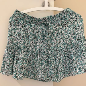 Blommig kortkjol med volang - Supersöt kortkjol med blommigt mönster i turkos, svart och vit. Kjolen har volang nertill och knytband i midjan för extra detalj. Perfekt för varma dagar och ger en fräsch look.