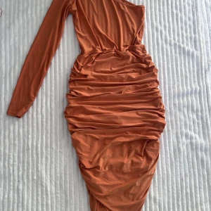 Rustfärgad one shoulder klänning från Boohoo - Säljer en snygg rostfärgad klänning från Boohoo med one shoulder-design och lång ärm. Klänningen har draperade detaljer längs sidorna och är tillverkad i ett mjukt, stretchigt material. Perfekt för dig som vill sticka ut!
