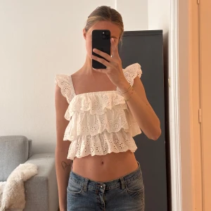 Vit volangtopp med broderade detaljer - Säljer en söt vit topp med volanger och broderade mönster av blommor. Toppen har breda axelband och är ärmlös med en luftig och romantisk känsla. Perfekt till jeans eller kjol för en somrig look!🥰storlek S men mer som en xs.