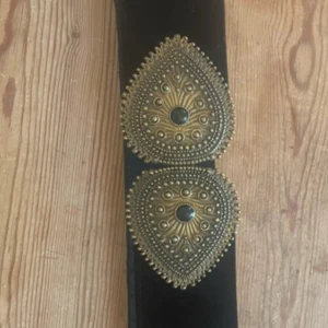 Svart sammetsbälte med stora guldspännen från Sezane - Unikt svart sammetsbälte från Sezane med två stora, dekorativa guldspännen med detaljerade mönster. Bältet är brett och har en lyxig känsla, perfekt för att lyfta en outfit med en statement-accessoar.