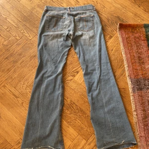 Blå bootcut jeans från Fornarina - Säljer ett par klassiska blå jeans från Fornarina med bootcut passform. Jeansen har slitna detaljer, råa kanter nertill och snygga bakfickor med dekorativa sömmar. Perfekta för dig som gillar en avslappnad och tidlös stil. Gylfen går ej att stänga men det tänker man inte på om man har en stickad tröja på sig eller längre topp!