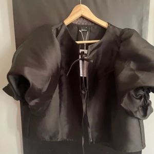 Gina tricot Svart puffärmad blus - Säljer en svart blus från Envii med stora puffärmar och rund halsringning. Blusen har en kortare passform och knytning framtill. Perfekt för dig som vill ha en statement-topp med volym och stil.storlek Xl passar large,nyskick med lappar 