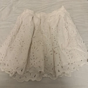 Vit kjol med broderade detaljer - Säljer en söt vit kjol med broderade blommönster och vågig kant. Kjolen har dragkedja bak och är fodrad. Perfekt för sommarens alla tillfällen!