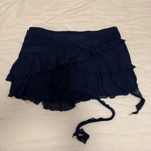 Mörkblå minikjol med volanger - Supersöt mörkblå minikjol med omlottmodell och volangdetaljer. Det är inbyggda shorts i kjolen! Kjolen har knytband i midjan och är tillverkad i ett lätt, flowy material som ger en luftig känsla. Perfekt för dig som gillar en feminin och trendig stil.