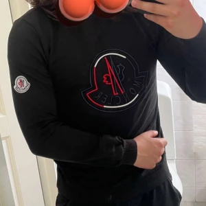 Svart sweatshirt från Moncler - Svart sweatshirt från Moncler med stort broderat logomärke på bröstet i rött, vitt och svart. Tröjan har rund halsringning, långa ärmar och ribbade muddar. Moncler-logga även på vänster ärm. Perfekt för en stilren och sportig look.