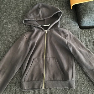 Hoodie med dragkedja XS - En stilren svart men lite sliten hoodie från 157 i storlek XS, ser mer svart ut i verkligheten. Har huva, dragkedja framtill och två stora fickor. Perfekt för lager-på-lager och chill dagar. Tillverkad i mjuk bomullsmix för skön känsla och enkel passform.