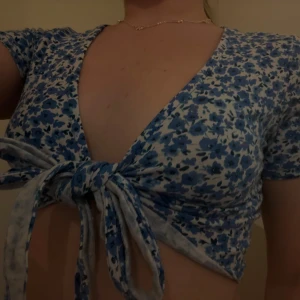 Blommig knytblus i blått och vitt - Supersöt blommig topp med knyt framtill i blått och vitt. Djup v-ringning och korta ärmar ger en fräsch och somrig vibe. Perfekt för dig som vill sticka ut med en feminin och trendig look.