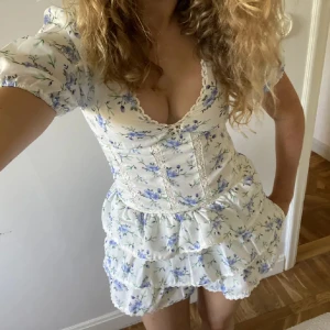 Blommig kortklänning med volang och puffärm - Supersöt vit kortklänning med blått blommönster, v-ringning och volangdetaljer. Klänningen har puffärmar och spets längs urringningen. Perfekt för sommarens alla tillfällen! Är köpt på Plick men säljer vidare då den ej kommer till användning är i storlek L men skulle säga storlek S/M är verkligen liten I storleken