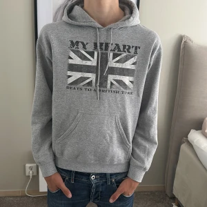 Grå hoodie med brittisk flagga - Grå hoodie med tryck av brittiska flaggan och texten 'MY HEART BEATS TO A BRITISH TUNE' på bröstet. Klassisk känguruficka framtill, snörning i huvan och mjukt material. Perfekt för dig som gillar streetstyle och vill ha en chill look.