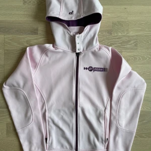 Ljusrosa kofta från Peak Performance - Ljusrosa kofta från Peak Performance med huva, dragkedja och snygga detaljer i lila. Jackan har broderad logga på bröstet och praktiska fickor med dragkedja. Perfekt för dig som gillar sportig och clean stil.