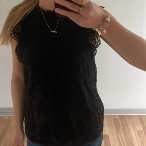 Svart spetsblus från Pieces - Supersnygg svart blus från Pieces med spetsdetaljer och broderade kanter vid ärmarna. Ärmlös modell med rund halsringning och knäppning bak i nacken. Perfekt att styla med jeans eller kjol för en trendig look.