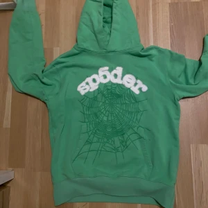 Grön Sp5der hoodie med spindelnät - Grön hoodie från Sp5der med stor vit logga och coolt spindelnätsmönster på framsidan. Tröjan har huva, magficka och långa ärmar. Perfekt statement-plagg för dig som gillar streetwear och unika prints.
