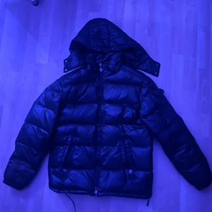 Svart pufferjacka med huva - Säljer en svart pufferjacka med huva och dragkedja framtill. Jackan har två stora fickor med dragkedja och en quiltad design som ger en riktigt fet look. Perfekt för kalla dagar när du vill hålla värmen och ändå se stilren ut.