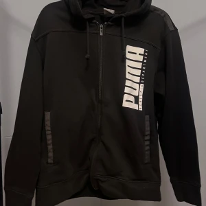 Svart hoodie från Puma med tryck - Svart hoodie från Puma med stor vit logga och text på bröstet. Tröjan har dragkedja, huva med snörning och två fickor framtill. Tillverkad i mjuk bomullsblandning, perfekt för en sportig och chill stil.