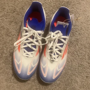 Adidas fotbollsskor i vitt och orange - Snygga Adidas fotbollsskor med vit bas, orangea och blå detaljer samt klassiska ränder på sidan. Skorna har snörning och plös i blått, samt dobbar i orange och vitt för bra grepp på planen. Perfekta för dig som vill sticka ut på matchen.