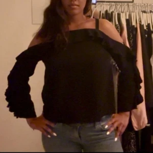 Svart offshoulder blus med volang - Säljer en svart offshoulder blus med långa ärmar och snygga volangdetaljer längs axlar och ärmar. Toppen har en rak passform och tunna axelband som ger en cool look. Perfekt för dig som vill sticka ut lite extra.