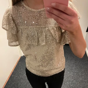 Glittrig topp med volangdetaljer - Säljer en glittrig topp i guld med volangdetaljer. Toppen har korta ärmar och en rund halsringning. Perfekt för en festlig kväll!