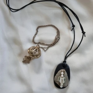 Två unika halsband med detaljer - Säljer två unika halsband. Det ena har ett intrikat silverfärgat hänge med filigranarbete och kedja. Det andra har ett svart snöre med ett hänge av sten, dekorerat med pärlor och ett hjärta i glas. Perfekt för dig som gillar unika smycken!