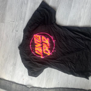 Svart t-shirt från Santa Cruz - Säljer en svart t-shirt från Santa Cruz med ett coolt tryck i neonrosa och gult på både fram- och baksidan. Perfekt för en avslappnad stil. T-shirten är kortärmad och gjord i ett bekvämt material. BEHÖVER STRYKAS