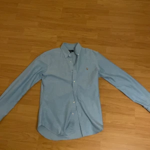 Ljusblå skjorta från Polo Ralph Lauren - Snygg ljusblå skjorta från Polo Ralph Lauren med klassisk krage och broderad logotyp på bröstet. Skjortan har långa ärmar och knappar framtill. Perfekt för en stilren look. 
