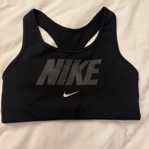 Svart sporttopp från Nike - Snygg svart sporttopp från Nike med Dri-FIT-teknologi. Toppen har ett stort Nike-logotyp i silver framtill och är perfekt för träning. Den är ärmlös och har en racerback-design för extra komfort och rörelsefrihet.